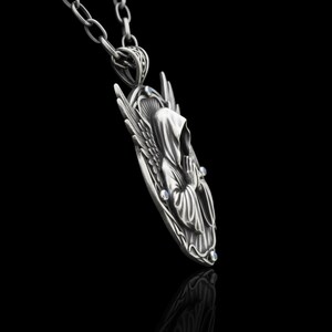 Puede incluir: Un collar con colgante plateado que representa una figura encapuchada con alas. El colgante presenta un dise&ntilde;o detallado de una figura rezando, adornada con peque&ntilde;as piedras preciosas azules. El collar cuelga de una cadena plateada sobre un fondo negro.