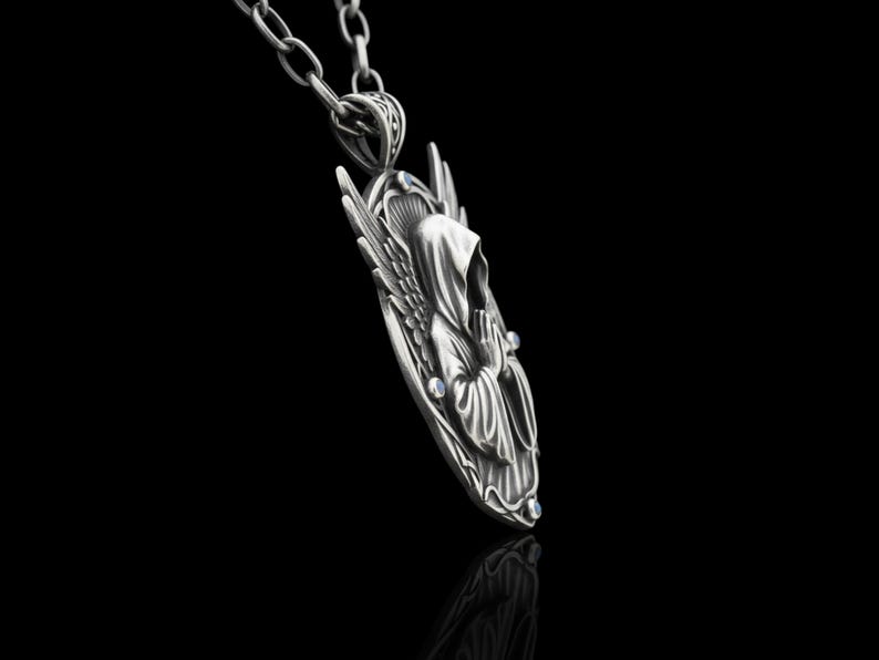 Puede incluir: Un collar con colgante plateado que representa una figura encapuchada con alas. El colgante presenta un dise&ntilde;o detallado de una figura rezando, adornada con peque&ntilde;as piedras preciosas azules. El collar cuelga de una cadena plateada sobre un fondo negro.