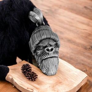 Puede incluir: Un colgante de plata con una escultura detallada de un gorila que lleva una boina de punto. El colgante está sobre una superficie de madera con una piña.