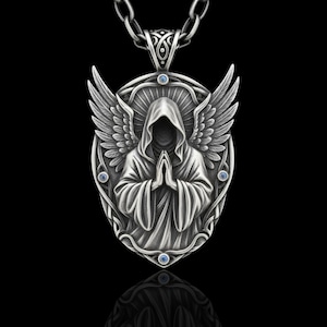 Puede incluir: Un collar con colgante de plata que representa una figura encapuchada con las manos juntas, alas de &aacute;ngel y detalles azules. El colgante ovalado cuelga de una cadena de plata. El dise&ntilde;o tiene detalles intrincados y un tema g&oacute;tico.