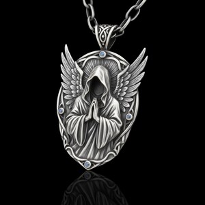 Puede incluir: Un collar con colgante plateado que presenta una figura encapuchada con las manos juntas, alas de &aacute;ngel y peque&ntilde;os detalles azules. El colgante ovalado cuelga de una cadena plateada, sobre un fondo negro.