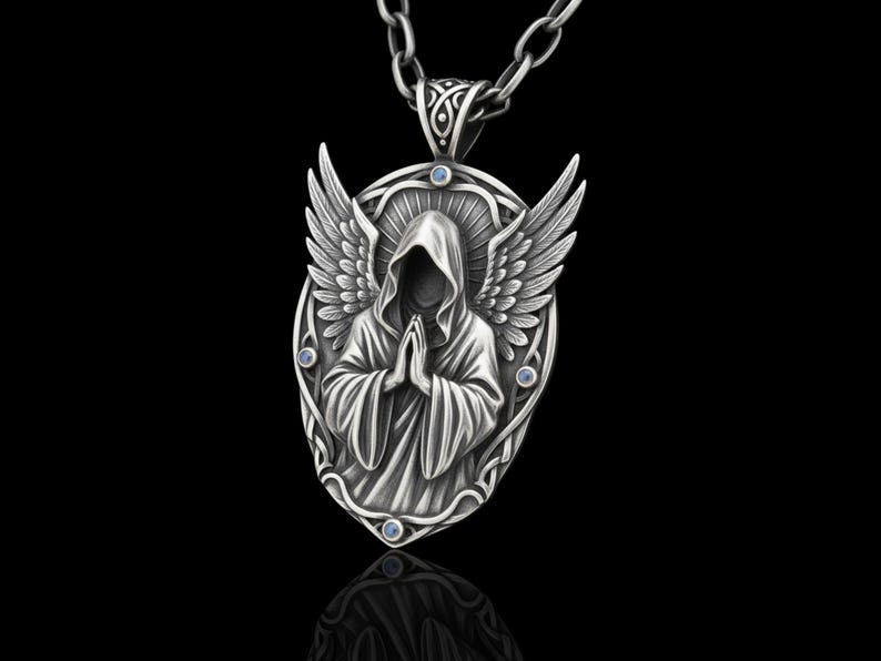 Puede incluir: Un collar con colgante plateado que presenta una figura encapuchada con las manos juntas, alas de &aacute;ngel y peque&ntilde;os detalles azules. El colgante ovalado cuelga de una cadena plateada, sobre un fondo negro.