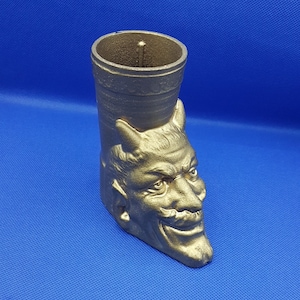 May include: A gold-colored devil-themed candle holder. The candle holder is shaped like a boot with the face of a devil on the front. The devil has horns, a mustache, and a sinister grin. The background is a solid blue.