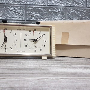 May include: Vintage chess clock with a cream-colored face and black hands. The clock has two separate dials, each with a set of numbers and a small second hand. The clock is in a rectangular frame with a beige box in the background.