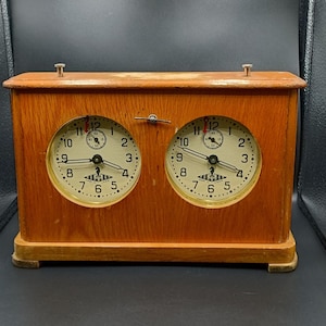 May include: A vintage wooden chess clock with two circular clock faces. The clocks have white faces with black numerals and hands, and a small lever on top. The wooden case is a light brown color.