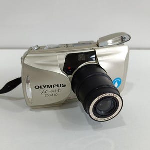 May include: A silver Olympus µ[mju:]-II Zoom 80 camera with a black lens. The camera has a built-in flash and a zoom lens with a 38-80mm range. The camera has a black strap attached.
