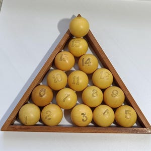 Könnte beinhalten: Ein Satz von fünfzehn nummerierten Billardkugeln, angeordnet in einem dreieckigen Holzrahmen. Die Kugeln sind hellgelb, jede mit einer schwarzen Nummer. Der Holzrahmen ist dunkelbraun.