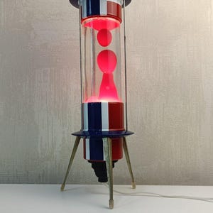 May include: A retro lava lamp with a red, white, and blue color scheme. The lamp has a cylindrical glass tube containing red lava, a top hat-shaped top, and a tripod base. The lamp is approximately 18 inches tall.