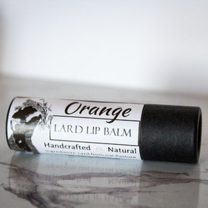 May include: A black tube of orange-scented lard lip balm with a white label that reads "Orange Lard Lip Balm Handcrafted Natural Ingredients Lard from our Pasture".