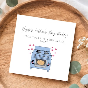 Pu&ograve; includere: Un biglietto bianco per la festa del pap&agrave; con il testo "Happy Father's Day Daddy!" e "From your little bun in the oven!". Presenta una grafica di forno blu con un panino all'interno e cuori rosa.