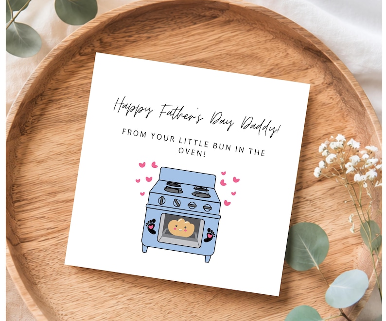 Pu&ograve; includere: Un biglietto bianco per la festa del pap&agrave; con il testo "Happy Father's Day Daddy!" e "From your little bun in the oven!". Presenta una grafica di forno blu con un panino all'interno e cuori rosa.