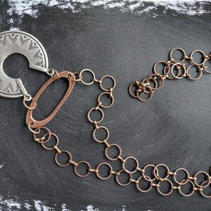 May include: A silver and copper necklace with a large, circular pendant featuring a geometric design. The pendant is attached to a chain of interlocking copper rings.