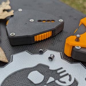 May include: Two black and orange 3D printed devices with a gear-like design. The devices are on a black surface with a white gear design.