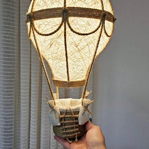Könnte beinhalten: Dekorative Heißluftballon-Lampe. Der Ballon besteht aus gewebtem Garn, mit einem Korb als Basis. Die Lampe strahlt ein warmes, einladendes Licht aus. Das Design integriert natürliche Materialien und Texturen, passend für ein Kinderzimmer.