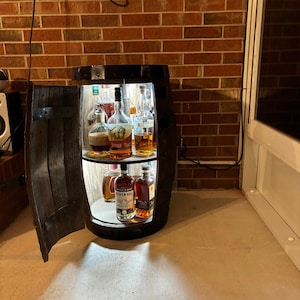 Op de afbeelding: Een rustieke drankkast van houten vat met een scharnierende deur, een verlicht interieur en twee planken met diverse drankflessen. De kast is gemaakt van donker gebeitst hout en heeft metalen banden. De binnenkant is verlicht met een zacht, warm licht.