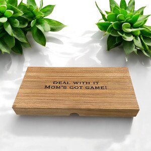 K&ouml;nnte beinhalten: Eine rechteckige Holzschachtel mit hellbrauner Maserung. Auf dem Deckel befindet sich der eingravierte schwarze Text "DEAL WITH IT MOM'S GOT GAME!", der mittig platziert ist. Die Schachtel hat eine kleine halbkreisf&ouml;rmige Aussparung vorne.