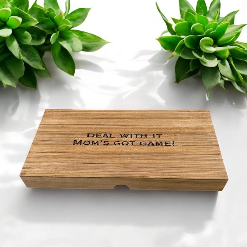K&ouml;nnte beinhalten: Eine rechteckige Holzschachtel mit hellbrauner Maserung. Auf dem Deckel befindet sich der eingravierte schwarze Text "DEAL WITH IT MOM'S GOT GAME!", der mittig platziert ist. Die Schachtel hat eine kleine halbkreisf&ouml;rmige Aussparung vorne.