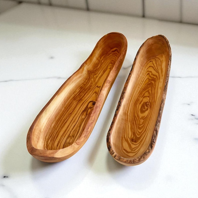 May include: Two elongated, narrow wooden bowls crafted from olive wood, featuring a smooth, polished interior surface that highlights the distinct, swirling brown and tan grain patterns and a natural, textured bark edge on the exterior sides.