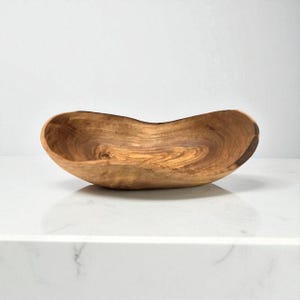 May include: An oval-shaped wooden bowl featuring a natural grain pattern with swirling light and dark brown tones. The bowl has an irregular, organic rim and a smooth, polished interior surface that highlights the unique texture of the wood.