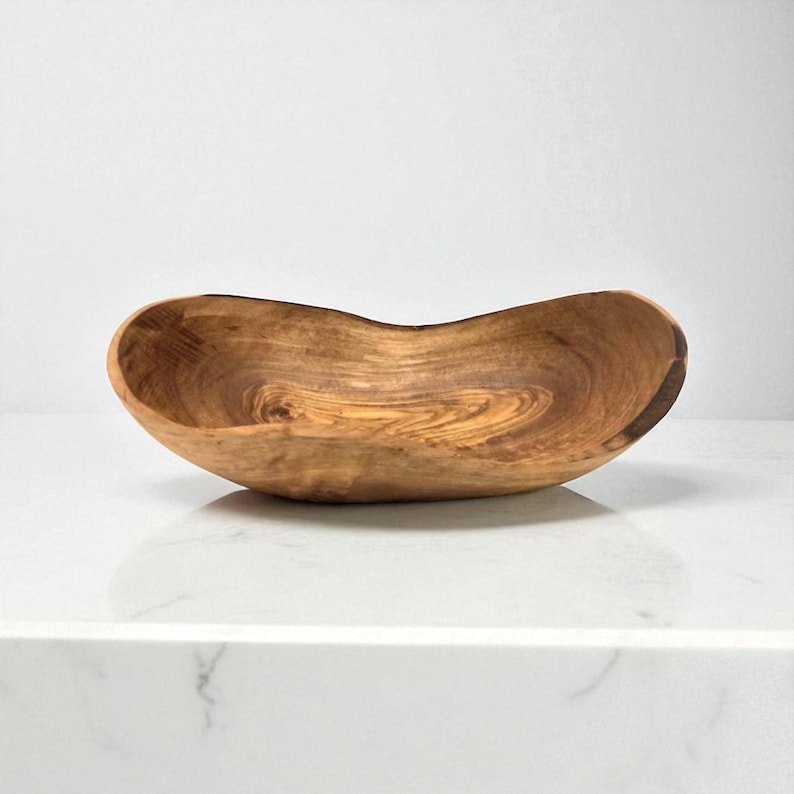 May include: An oval-shaped wooden bowl featuring a natural grain pattern with swirling light and dark brown tones. The bowl has an irregular, organic rim and a smooth, polished interior surface that highlights the unique texture of the wood.