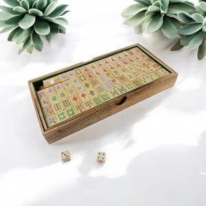 K&ouml;nnte beinhalten: Eine rechteckige Holzschachtel mit einem Satz Mahjong-Spielsteine, auf deren Oberfl&auml;chen verschiedene bunte chinesische Schriftzeichen und Symbole eingraviert sind. Zwei kleine quadratische W&uuml;rfel mit blauen und gr&uuml;nen Punkten liegen davor.