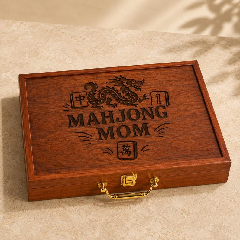K&ouml;nnte beinhalten: Eine rechteckige Holzbox mit dunkelbrauner Oberfl&auml;che sowie einem goldfarbenen Metallgriff und Verschluss. Der Deckel zeigt ein lasergraviertes Design mit einem Drachen, dekorativen Linien, chinesischen Schriftzeichen und dem Text "MAHJONG MOM".