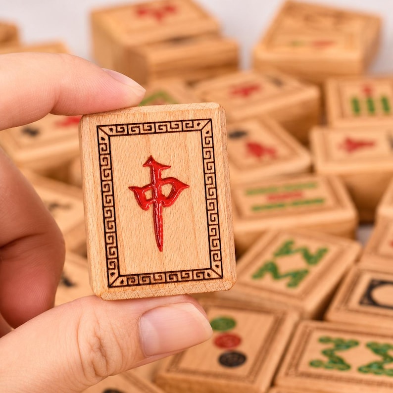 K&ouml;nnte beinhalten: Ein rechteckiger Mahjong-Spielstein aus Holz mit einem roten chinesischen Schriftzeichen in der Mitte, umgeben von einem dekorativen Rand. Der Spielstein ist hellbraun mit einer nat&uuml;rlichen Holzmaserung und liegt inmitten weiterer Spielsteine.