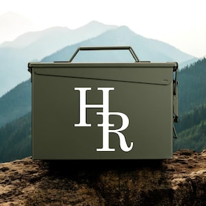 Puede incluir: Una caja de municiones de metal color verde oliva con un asa de transporte superior y una tapa con bisagras. El frente de la caja muestra las letras mayúsculas blancas H y R apiladas verticalmente en una fuente serif centrada.