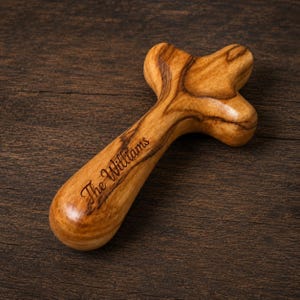 Könnte beinhalten: Ein glattes, poliertes Holzkreuz mit einer abgerundeten, ergonomischen Form, die zum Halten konzipiert ist. Das hellbraune Holz weist durchgehend dunkle, wirbelnde Maserungen auf. Der Text "The Williams" ist in dunkler Schreibschrift eingraviert.