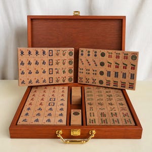 K&ouml;nnte beinhalten: Ein Mahjong-Spiel aus Holz in einem braunen Koffer mit goldenem Metallverschluss und Griff. Das Set enth&auml;lt zahlreiche rechteckige Spielsteine mit eingravierten chinesischen Schriftzeichen, Blumenmotiven und Joker-Symbolen in Schwarz, Rot und Gr&uuml;n.