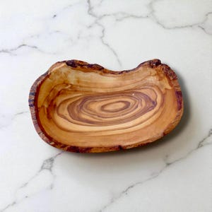 May include: A shallow, oval-shaped wooden bowl crafted from olive wood, featuring a natural live edge and prominent, swirling brown grain patterns that create a distinct, organic appearance across the smooth, polished interior surface.