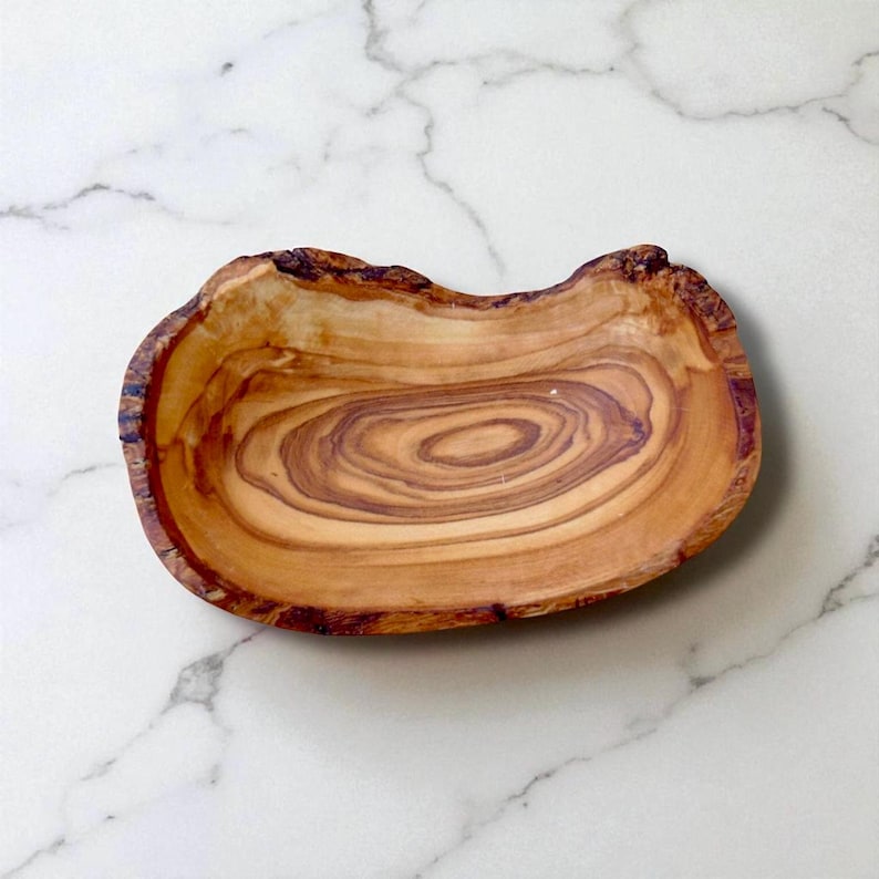 May include: A shallow, oval-shaped wooden bowl crafted from olive wood, featuring a natural live edge and prominent, swirling brown grain patterns that create a distinct, organic appearance across the smooth, polished interior surface.