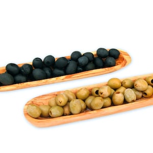 May include: Two elongated, oval-shaped wooden serving dishes with a natural grain pattern. One dish is filled with dark black olives, while the other dish is filled with light green olives. The dishes are positioned diagonally against a white background.