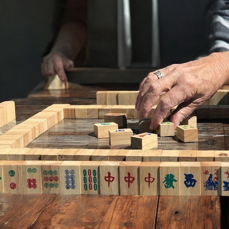 K&ouml;nnte beinhalten: Ein Set aus h&ouml;lzernen Mahjong-Spielsteinen auf einer dunklen Holzoberfl&auml;che, verziert mit verschiedenen eingravierten Symbolen und Schriftzeichen in roter, gr&uuml;ner und blauer Tinte, teils aufrecht in einer Reihe, teils flach in der Mitte.