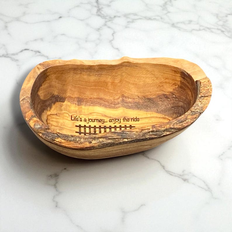 May include: An oval-shaped wooden bowl with a natural bark edge and visible wood grain. The interior is engraved with the text "Life's a journey... enjoy the ride" positioned above a graphic illustration of a railroad track.