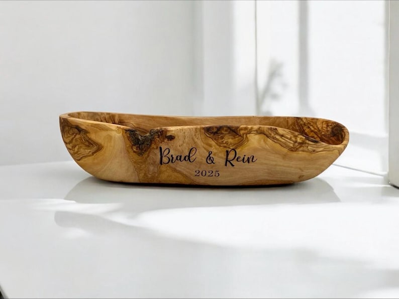 May include: An elongated, natural-edged wooden bowl crafted from olive wood with a distinct grain pattern. The exterior is engraved with the text "Brad & Rein" above the year "2025" in a dark, script-style font centered on the side.