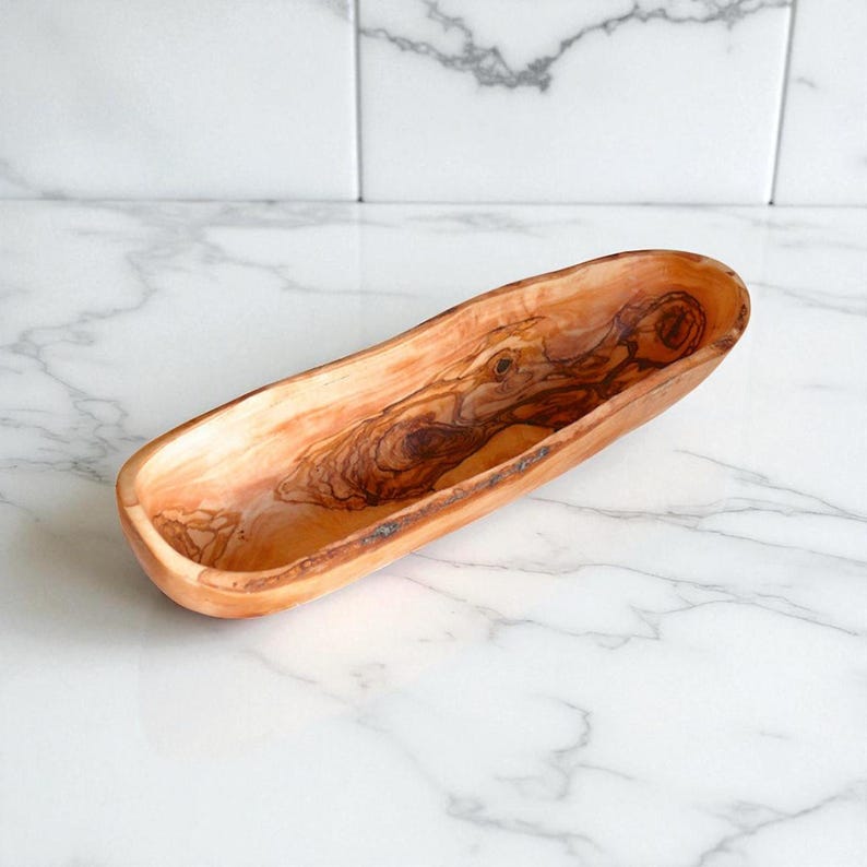 May include: An elongated, oval-shaped wooden bowl crafted from olive wood, featuring a smooth, polished finish that highlights the natural, swirling grain patterns and rich, warm brown and golden tones characteristic of the wood's organic texture.