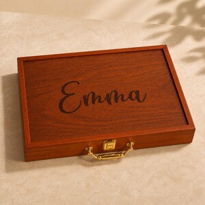 K&ouml;nnte beinhalten: Eine rechteckige Aufbewahrungsbox aus Holz mit dunkelbrauner Holzmaserung. Auf dem Deckel ist der Name Emma in Schreibschrift eingraviert. Die Box verf&uuml;gt &uuml;ber einen goldfarbenen Metallverschluss und einen passenden goldfarbenen Metallgriff.
