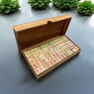 K&ouml;nnte beinhalten: Ein Mahjong-Spiel aus Holz in einer rechteckigen Holzschatulle mit offenem Deckel. Das Set enth&auml;lt zahlreiche rechteckige Spielsteine mit eingravierten chinesischen Schriftzeichen und Symbolen in roter, blauer und gr&uuml;ner Tinte, ordentlich in Reihen.
