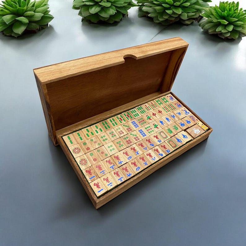 K&ouml;nnte beinhalten: Ein Mahjong-Spiel aus Holz in einer rechteckigen Holzschatulle mit offenem Deckel. Das Set enth&auml;lt zahlreiche rechteckige Spielsteine mit eingravierten chinesischen Schriftzeichen und Symbolen in roter, blauer und gr&uuml;ner Tinte, ordentlich in Reihen.
