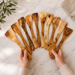 May include: A collection of nine wooden spatulas arranged in a fan shape. Each spatula is crafted from light brown wood with distinct dark brown grain patterns and knots, featuring a flat, wide head and a smooth, tapered handle for kitchen use.