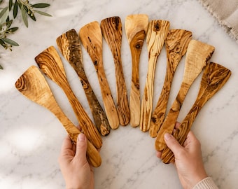 May include: A collection of nine wooden spatulas arranged in a fan shape. Each spatula is crafted from light brown wood with distinct dark brown grain patterns and knots, featuring a flat, wide head and a smooth, tapered handle for kitchen use.