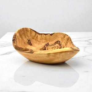 May include: A small, hand-carved wooden bowl made from olive wood, featuring a natural, irregular live edge and a distinct, swirling grain pattern in shades of light tan and dark brown. The bowl has a smooth, polished interior surface.