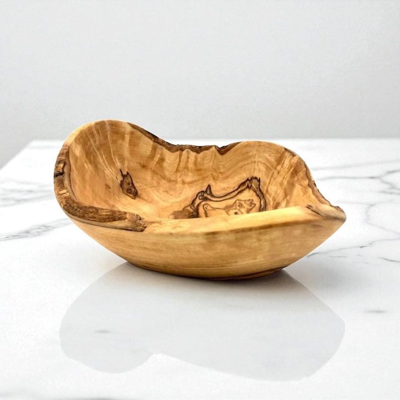May include: A small, hand-carved wooden bowl made from olive wood, featuring a natural, irregular live edge and a distinct, swirling grain pattern in shades of light tan and dark brown. The bowl has a smooth, polished interior surface.