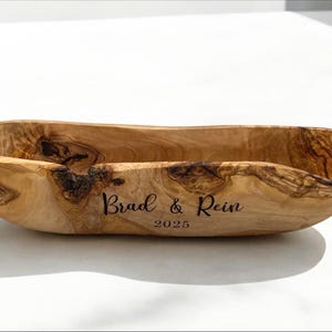 May include: An elongated, oval-shaped wooden bowl with a natural grain pattern and irregular, organic edges. The exterior is engraved with the text "Brad & Rein" above the year "2025" in a dark, script-style font centered on the side.