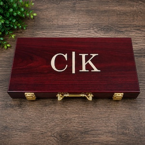 May include: A rectangular dark cherry wood box with a smooth finish, featuring the engraved letters C and K separated by a vertical line in the center. The box includes two gold-colored metal latches and a central gold-colored metal handle.