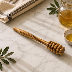 May include: A wooden honey dipper with a natural grain pattern and the text The Williams engraved on the handle. The dipper features a long, slender stem and a grooved head designed for drizzling honey, showcasing a smooth, polished finish.