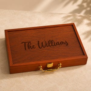 K&ouml;nnte beinhalten: Eine rechteckige Aufbewahrungsbox aus Holz mit dunkelbrauner Holzmaserung. Auf dem Deckel befindet sich der eingravierte Schriftzug The Williams in Schreibschrift. Die Box verf&uuml;gt &uuml;ber einen goldfarbenen Metallverschluss und einen passenden Metallgriff.