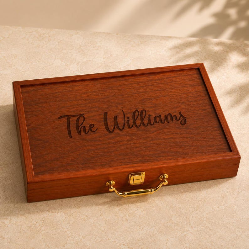 K&ouml;nnte beinhalten: Eine rechteckige Aufbewahrungsbox aus Holz mit dunkelbrauner Holzmaserung. Auf dem Deckel befindet sich der eingravierte Schriftzug The Williams in Schreibschrift. Die Box verf&uuml;gt &uuml;ber einen goldfarbenen Metallverschluss und einen passenden Metallgriff.