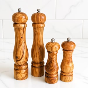 May include: Four wooden pepper mills with a distinct natural grain pattern and a light brown color. Each mill features a rounded top, a curved body, and a small metallic knob at the very peak. The set includes two taller mills and two shorter mills.
